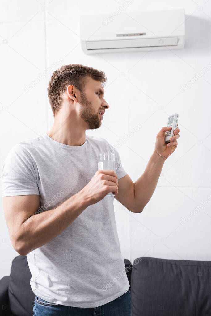 sad man holding glass of water while trying to switch on air conditioner with broken remote controller at home 