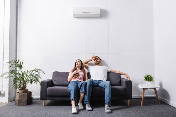 young sad couple suffering from heat while sitting home with broken air conditioner 