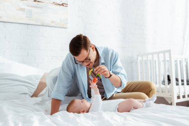 Mutlu genç adam bebek çıngırağıyla oynuyor. Yatakta uzanmış tatlı bir bebek var.