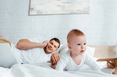 Akıllı telefondan konuşan bir adamın yatakta sürünen şirin oğluna dokunuşunun seçici odağı.