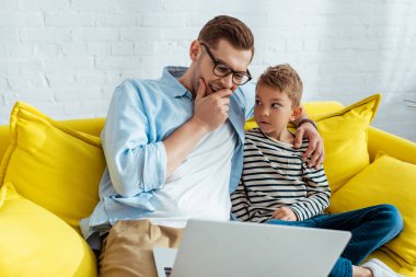 Genç baba kanepede oturup dizüstü bilgisayar kullanırken gülüyor ve sarılıyor.