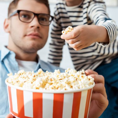 Oğlanın kovadan patlamış mısır alırken dikkatli bir babanın televizyon izlediği görüntüler.