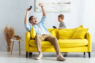 KYIV, UKRAINE - JUNE 9, 2020: excited man showing winner gesture while playing video game with son clipart