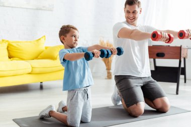 Gülümseyen baba ve oğlu halterlerle egzersiz yaparken spor yaparken dizlerinin üstünde spor paspası üzerinde duruyorlardı.