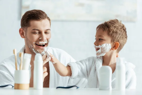 有选择地专注于微笑男孩在快乐的父亲脸上涂上剃须泡沫 — 图库照片