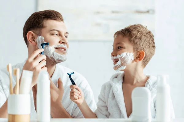 满脸泡沫的男孩看着刮胡子的父亲 选择性地专注于微笑 — 图库照片