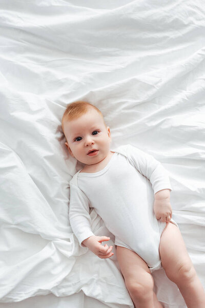 top view of adorable little boy in romper lying on bedding and looking at camera