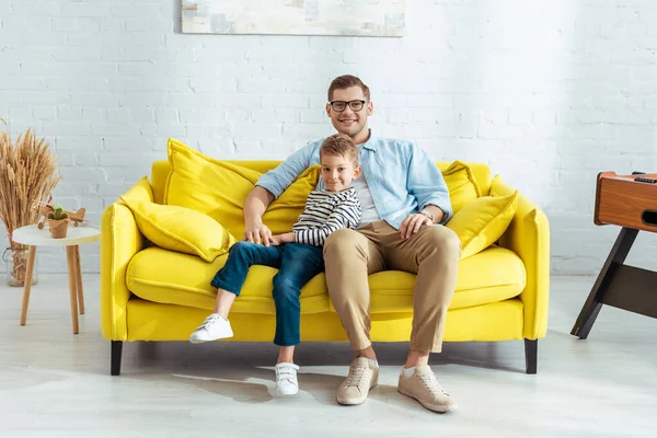 快乐的父亲和儿子坐在黄色的沙发上看着相机 — 图库照片