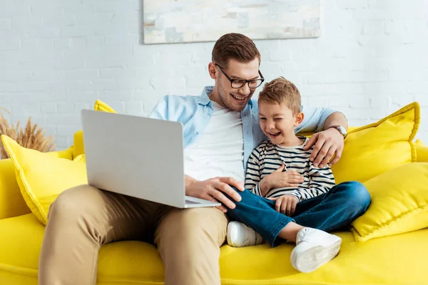 坐在黄色沙发上 带着笔记本电脑 兴奋的父亲和儿子笑了 — 图库照片