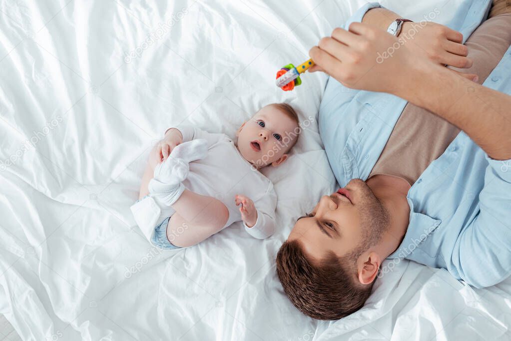 top view of young father playing with baby rattle over cute little son lying in bed