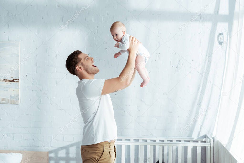 excited young father holding adorable baby boy in outstretched hands near baby cot