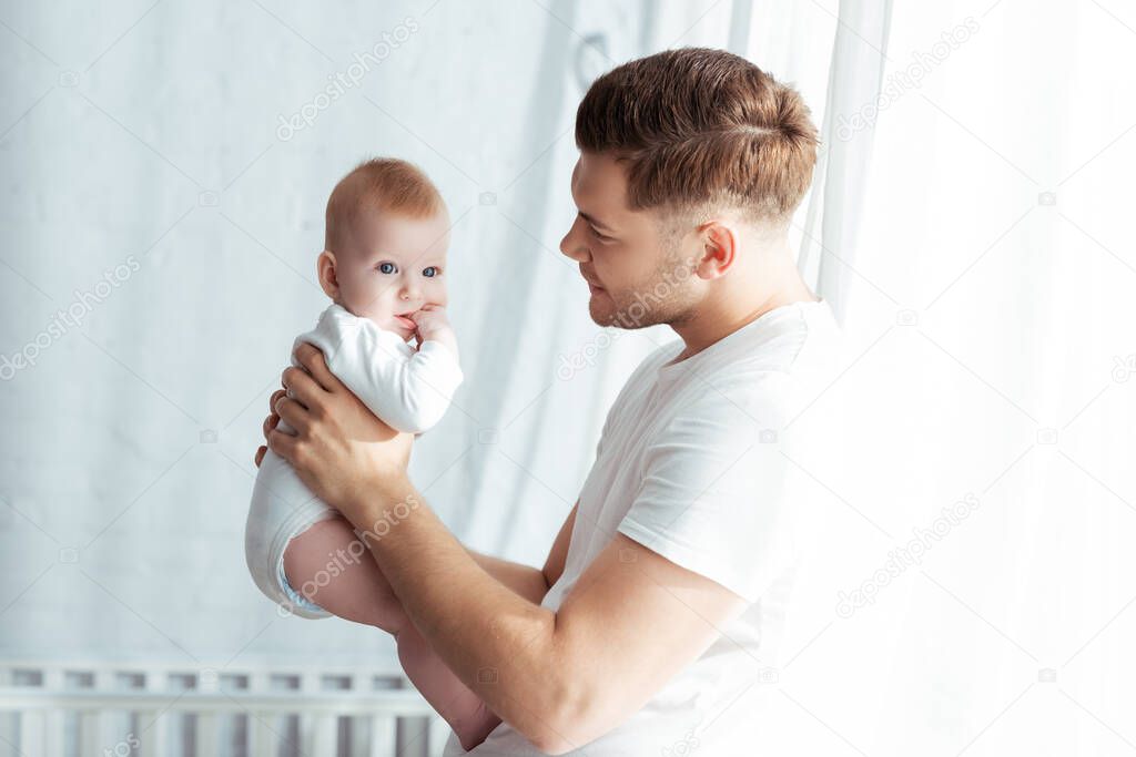 young father looking at adorable baby boy while holding him in bedroom