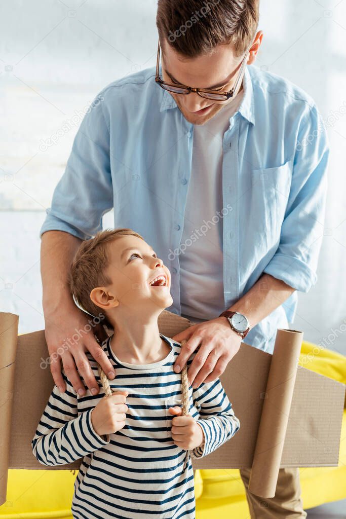 smiling father touching shoulders of happy son with cardboard plane wings on back