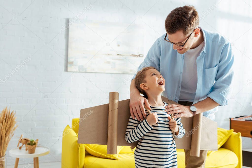 smiling father touching shoulders of adorable son with cardboard plane wings on back