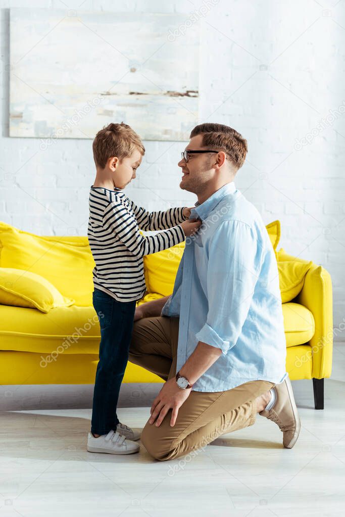 side view of adorable kid buttoning shirt of smiling father standing on knee