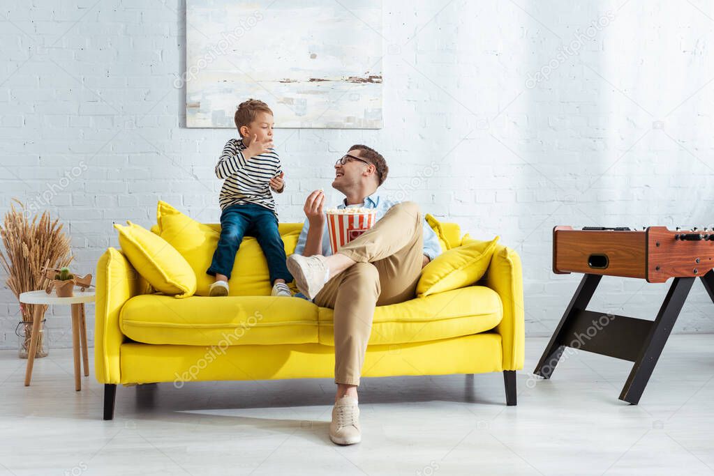 happy father and son eating popcorn while sitting on yellow sofa