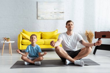 father and son sitting in lotus poses on fitness mat with closed eyes clipart