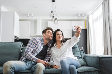 Gülümseyen kadın akıllı telefonuyla selfie çekiyor. Evdeki kanepede erkek arkadaşının yanında. 