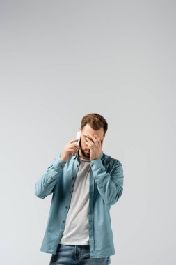 confused young man talking on smartphone and showing facepalm isolated on grey clipart