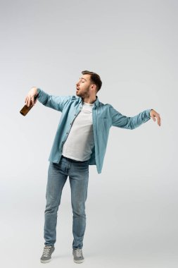 Genç adam, kukla gibi poz veriyor. Gri üzerinde bira şişesi var.