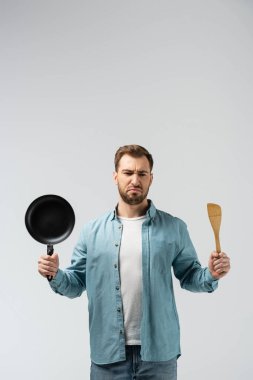 displeased young man with frying pan and spatula isolated on grey clipart