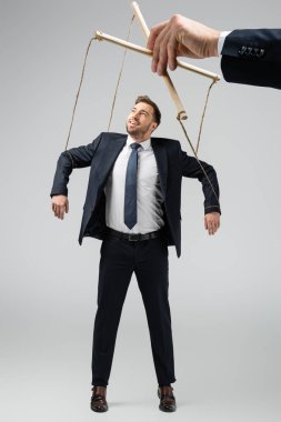 cropped view of puppeteer holding smiling businessman marionette on strings isolated on grey clipart