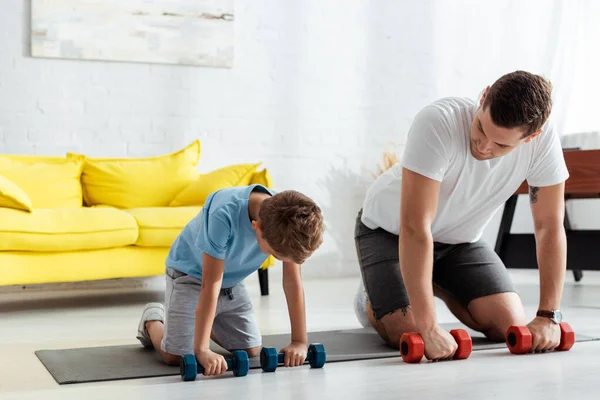 有可爱儿子的年轻父亲带着哑铃在健身垫上锻炼 — 图库照片