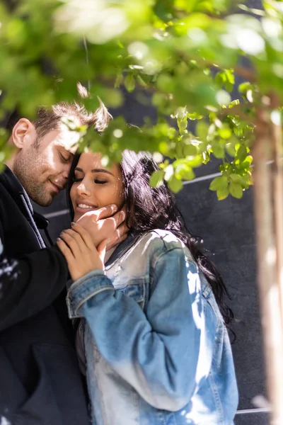 Selective focus of man touching face of smiling girlfriend near tree outdoors