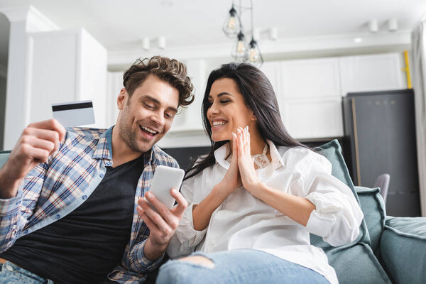 Selective focus of smiling man holding credit card and smartphone near girlfriend showing please gesture at home 