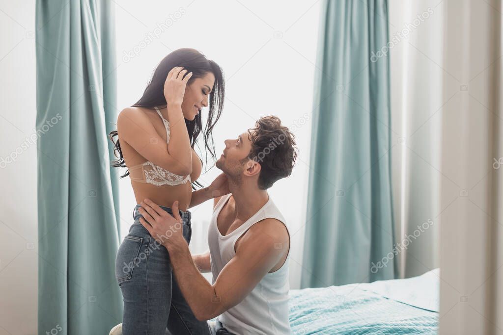 Side view of handsome man embracing smiling woman in bra in bedroom