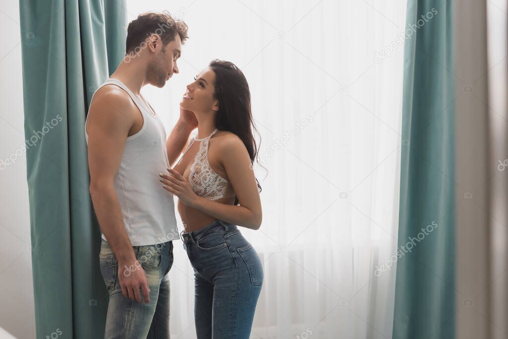 Side view of handsome man touching face of smiling girlfriend in bra and jeans near window 
