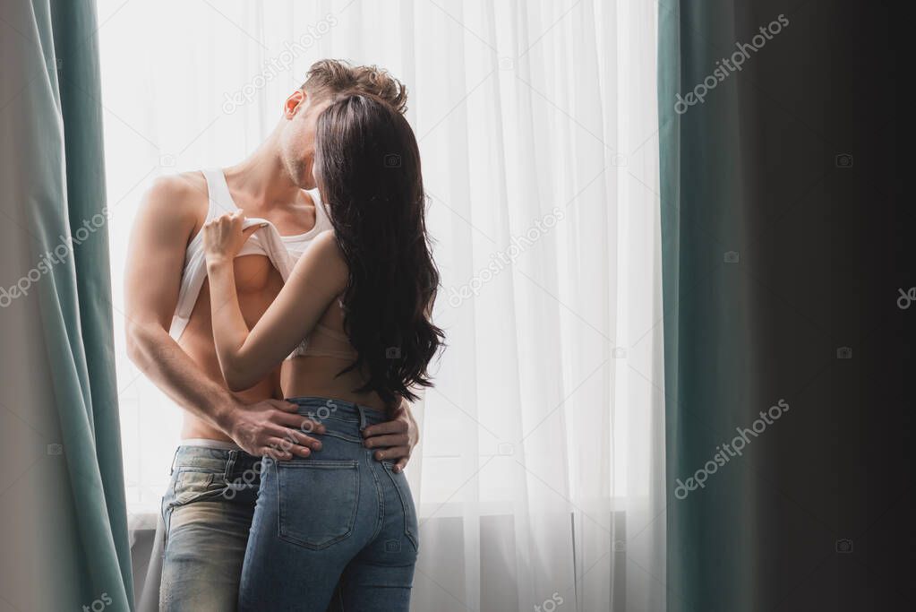 Selective focus of sexy woman taking off sleeveless shirt from muscular boyfriend while kissing near window 