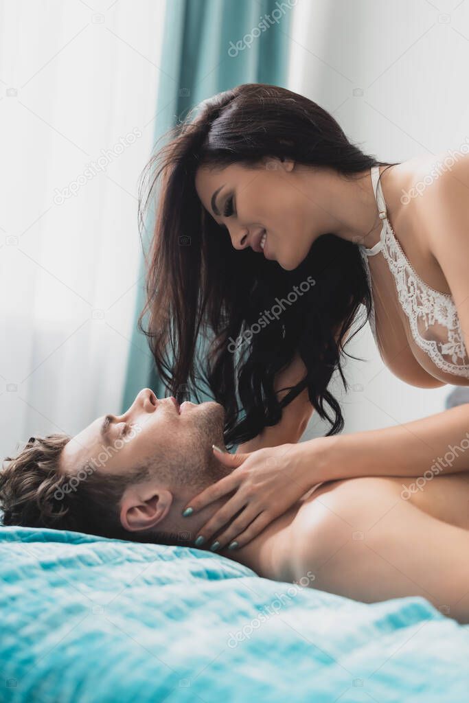 Side view of sensual smiling woman touching neck of muscular boyfriend on bed 