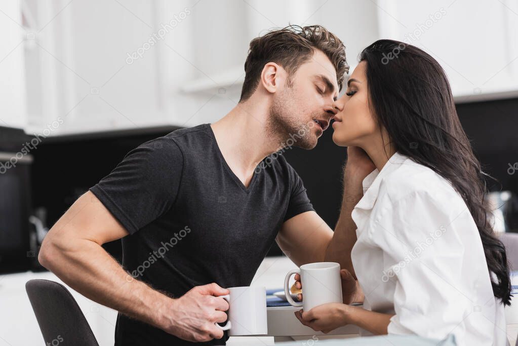 Handsome man kissing beautiful girlfriend while drinking coffee in kitchen 