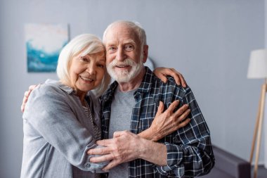 happy senior couple smiling and embracing while looking at camera clipart