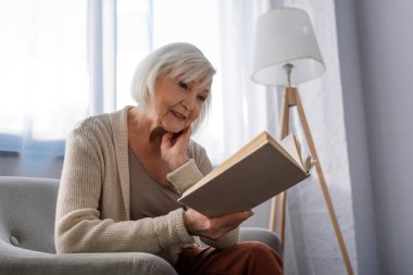 Pozitif kıdemli kadın koltukta oturup kitap okurken gülümsüyor.