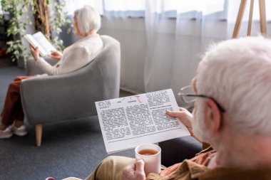 Karısı koltukta kitap okurken, yaşlı bir adamın elinde bir fincan çay tutup gazete okuduğu seçici bir odak noktası.