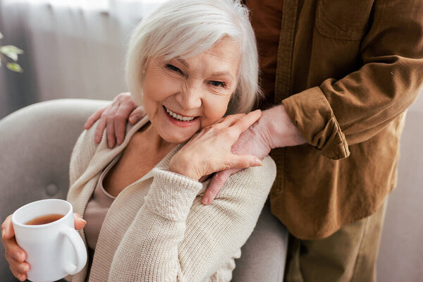 cropped view of man touching shoulders of cheerful senior wife holding cup of tea and smiling at camera