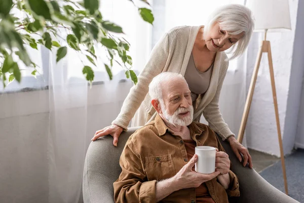 Senior Woman Standing Senior Husband Sitting Armchair Cup Tea — Stock Photo, Image