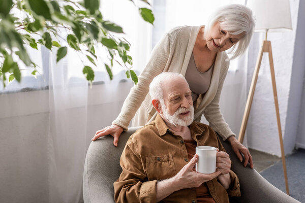 senior woman standing near senior husband sitting in armchair with cup of tea