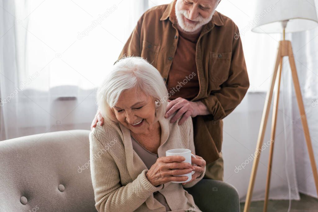 cheerful senior man touching shoulders of happy wife sitting in armchair with cup of tea