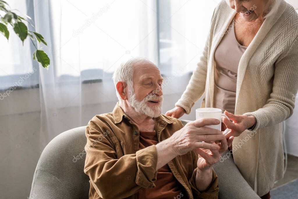 cropped view of senior woman giving cup of tea to smiling husband sitting in armchair