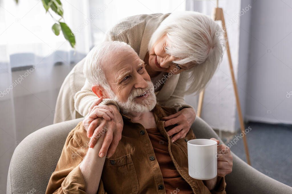 smiling woman hugging happy senior husband sitting with cup of tea