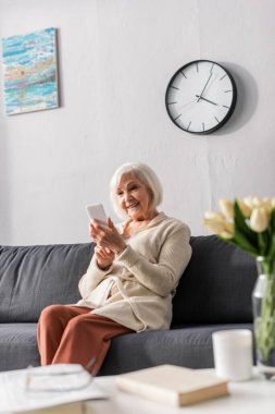 Kanepede otururken akıllı telefon kullanan mutlu yaşlı bir kadının seçici odağı. 