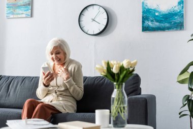 Heyecanlı son sınıf güzeli gülümserken masanın yanında lale buketiyle akıllı telefon kullanıyor.
