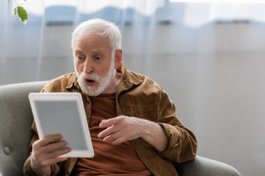 shocked senior man pointing with finger while using digital tablet clipart