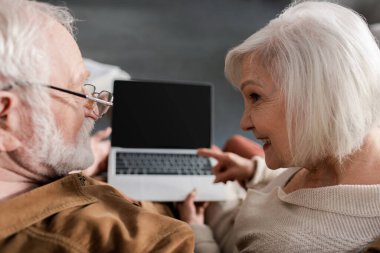 Kocanın yanında otururken dizüstü bilgisayarına boş ekranla bakan gülümseyen yaşlı kadının genel görünümü, seçici odaklanma