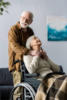 Gülümseyen kıdemli adam engelli karısının elini tutuyor tekerlekli sandalyede oturuyor. 