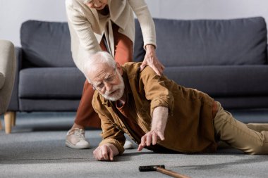 senior man lying on floor and trying to get walking stick while wife helping him clipart
