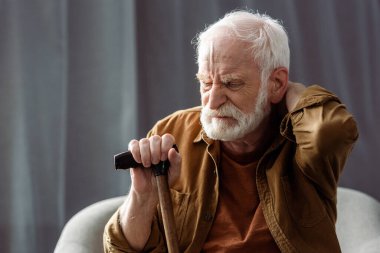 lonely senior man sitting with closed eyes, touching neck and holding walking stick  clipart